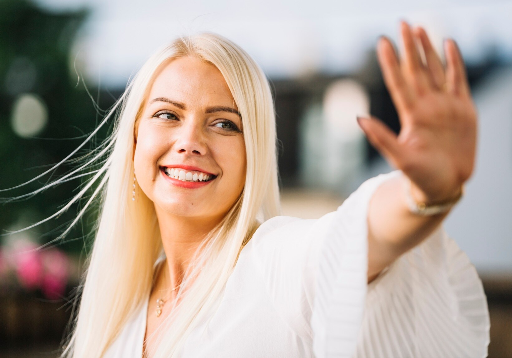 A confident woman with long blonde hair smiling brightly while waving, radiating self-confidence for women.